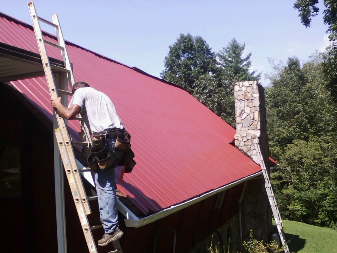 Roofer on a ladder beside a red metal roof and stone chimney — Soffit & Fascia Repair by Roofing Brothers in Red Oak